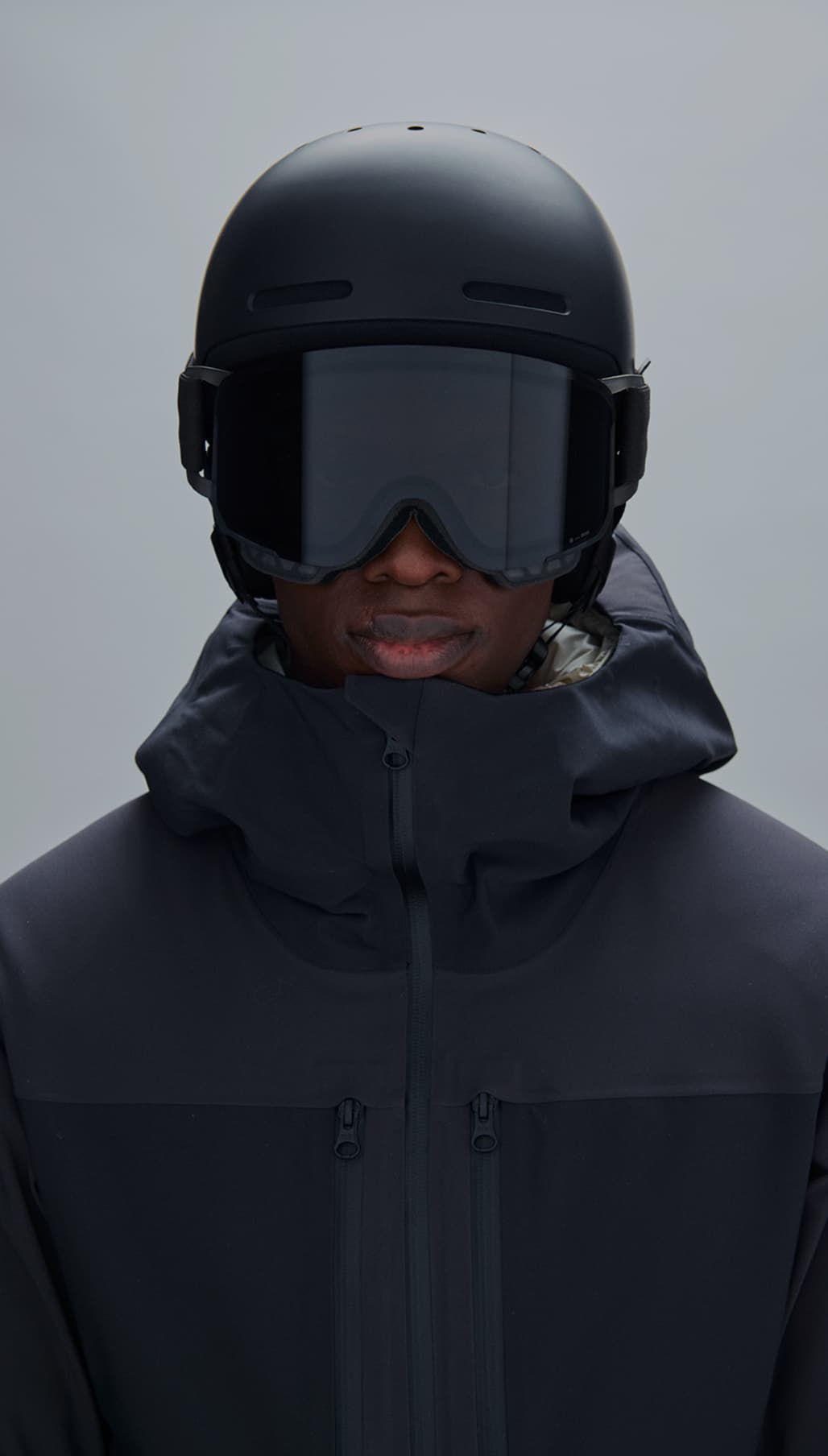 Person in a dark ski jacket, helmet, and goggles, facing forward against a gray background.
