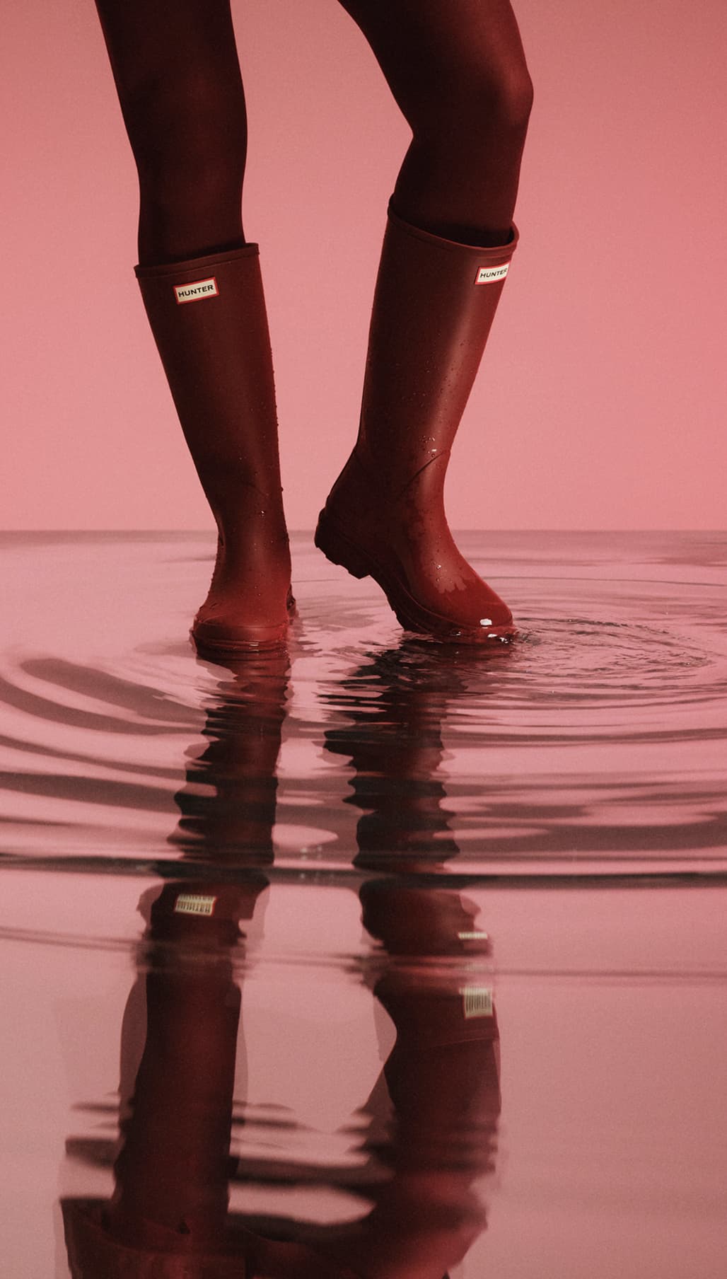 Person wearing red rain boots standing in shallow water, creating ripples, against a pink background.