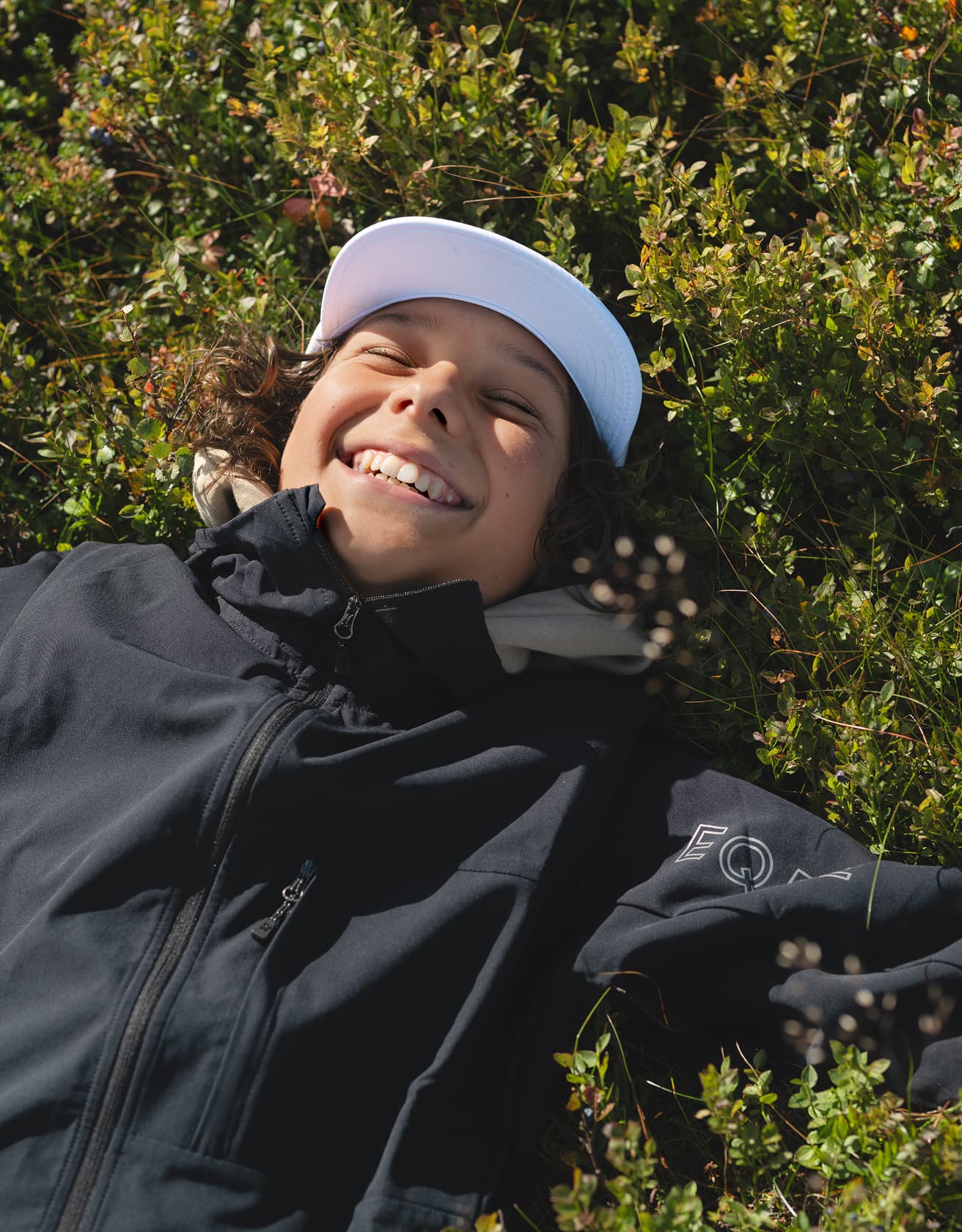 Smiling child in a white cap lies on greenery, wearing a dark jacket, enjoying a sunny day.