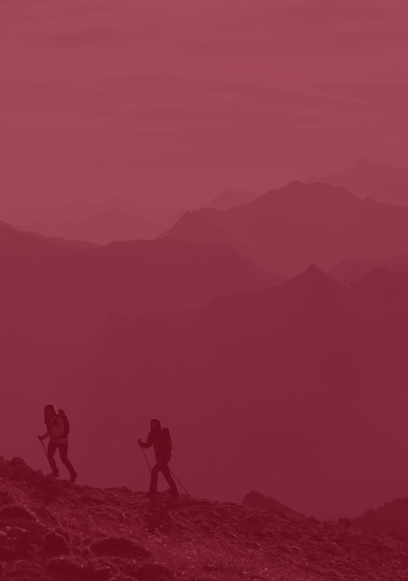 Two hikers traverse a mountain ridge, silhouetted against a hazy, red-tinted backdrop of distant peaks.