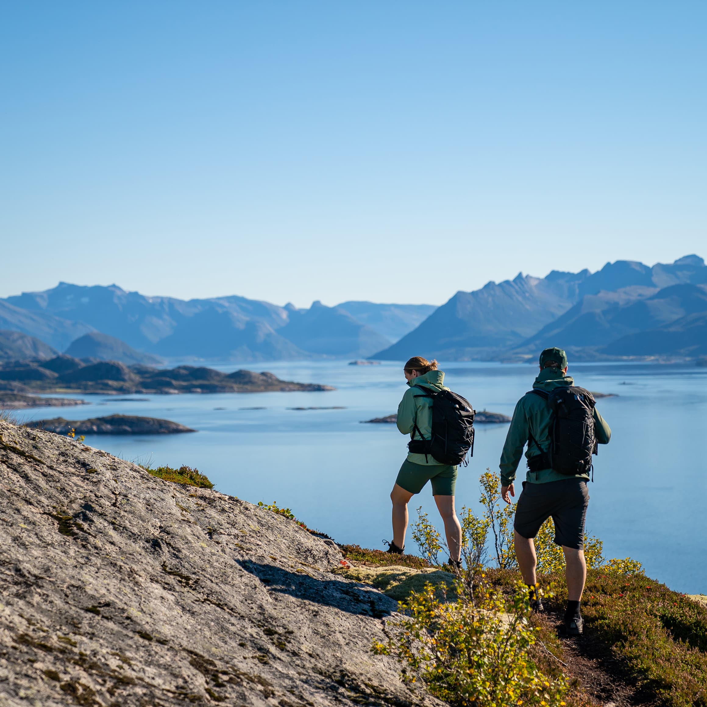 Two hikers walking on a rocky trail overlooking a scenic fjord and mountain landscape under a clear blue sky