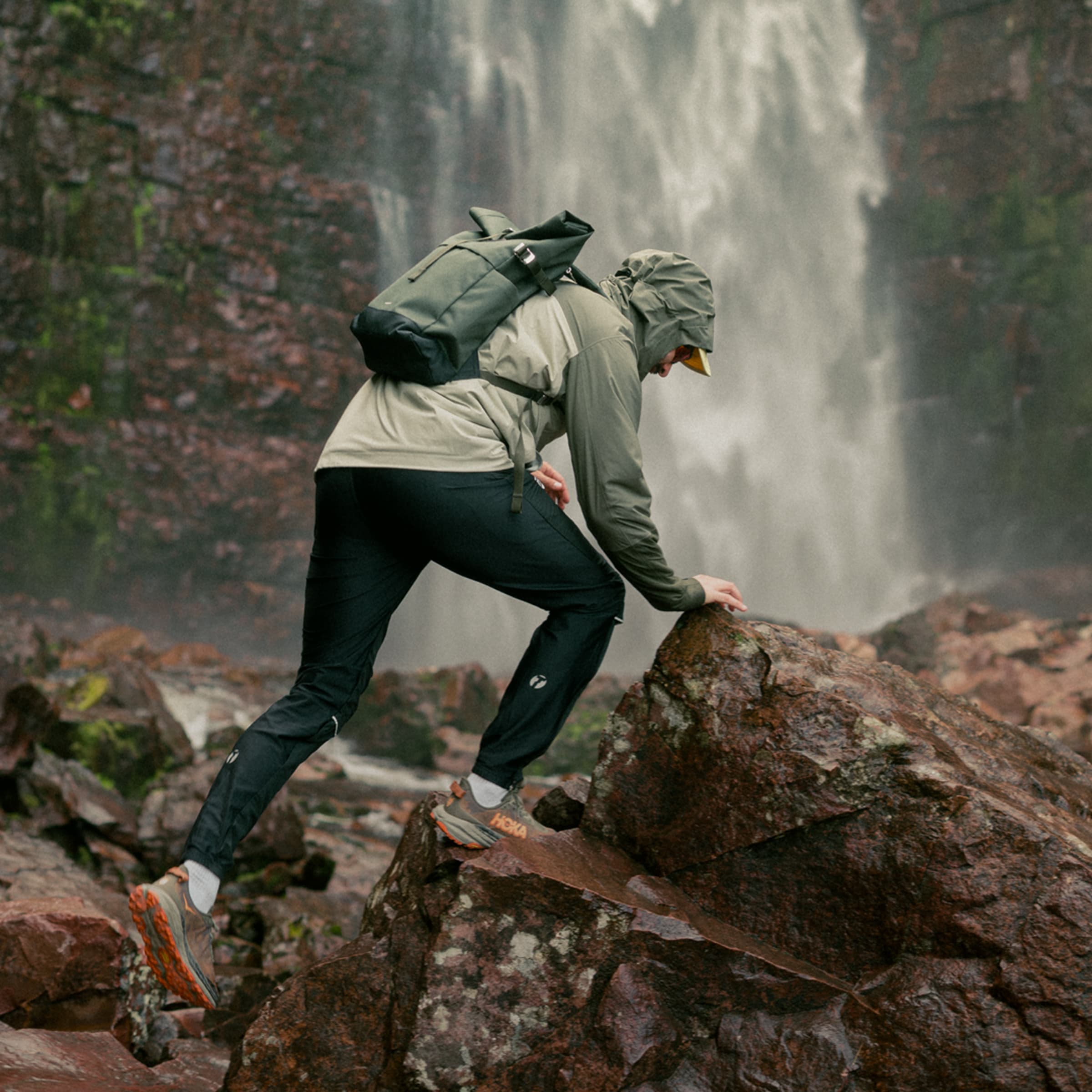 Person in outdoor gear climbing rocks near a waterfall, wearing a hooded jacket and backpack.