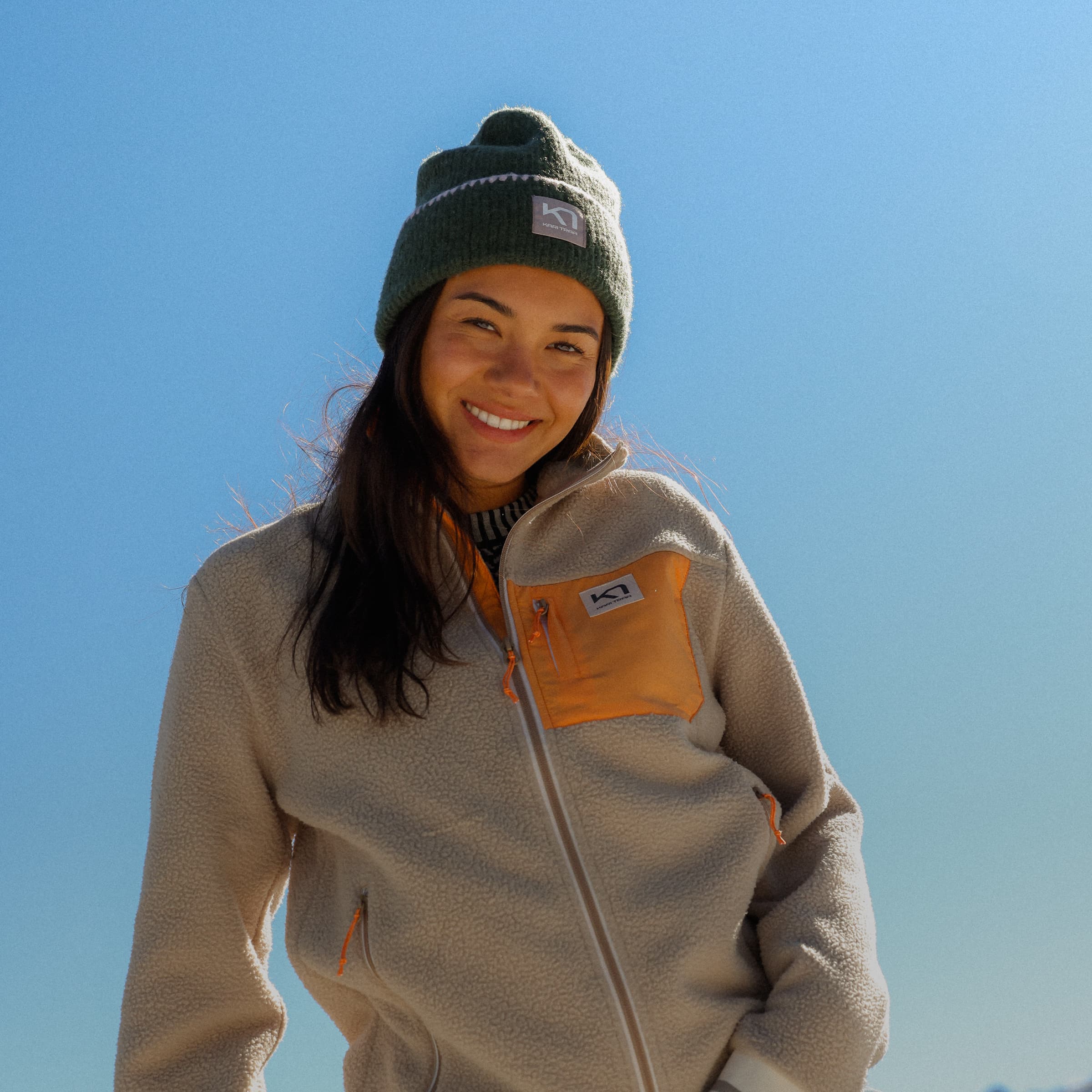 Woman smiling outdoors in a beige midlayer and green beanie.