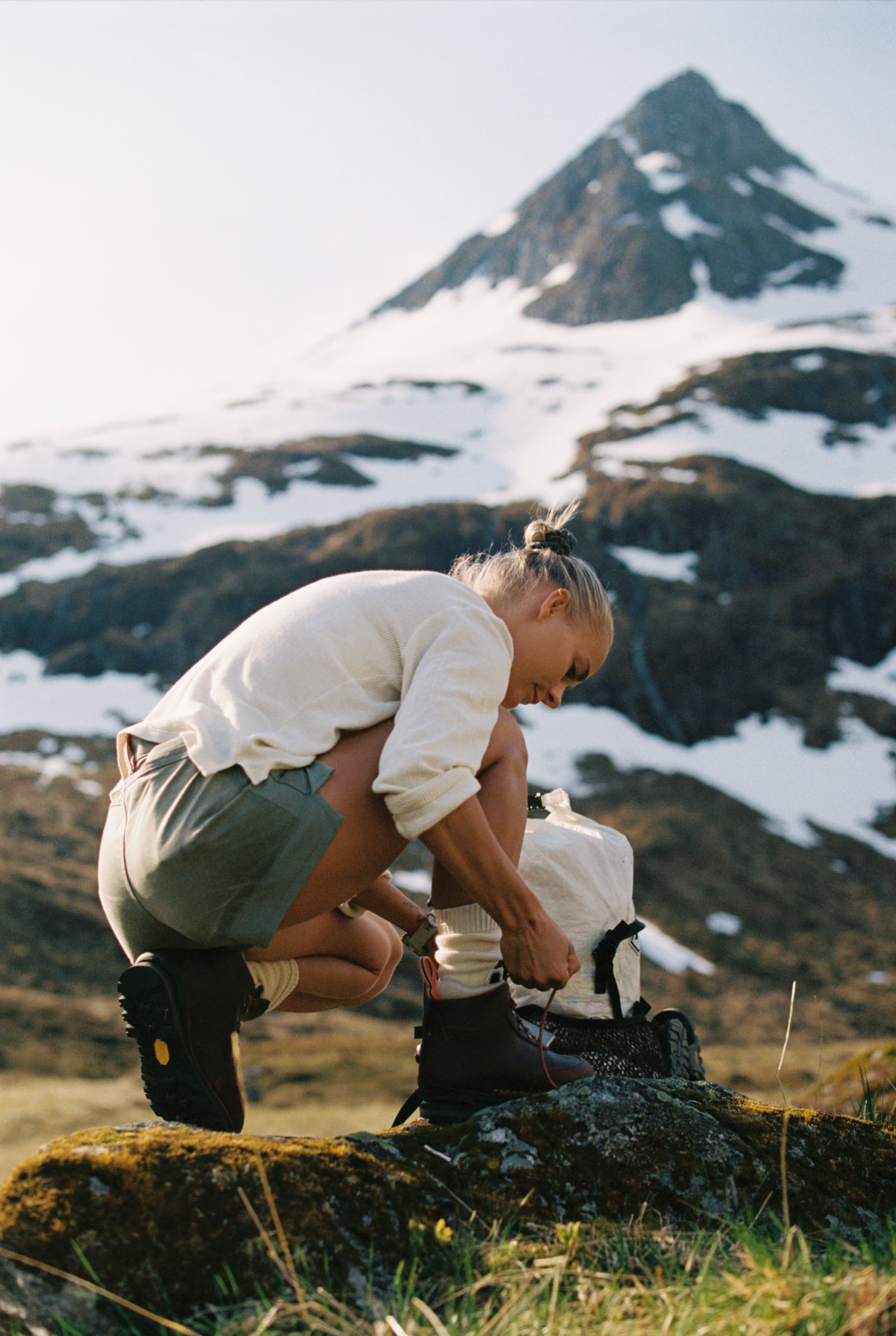 A person ties hiking boots on a mossy rock with a snow-capped mountain in the background.