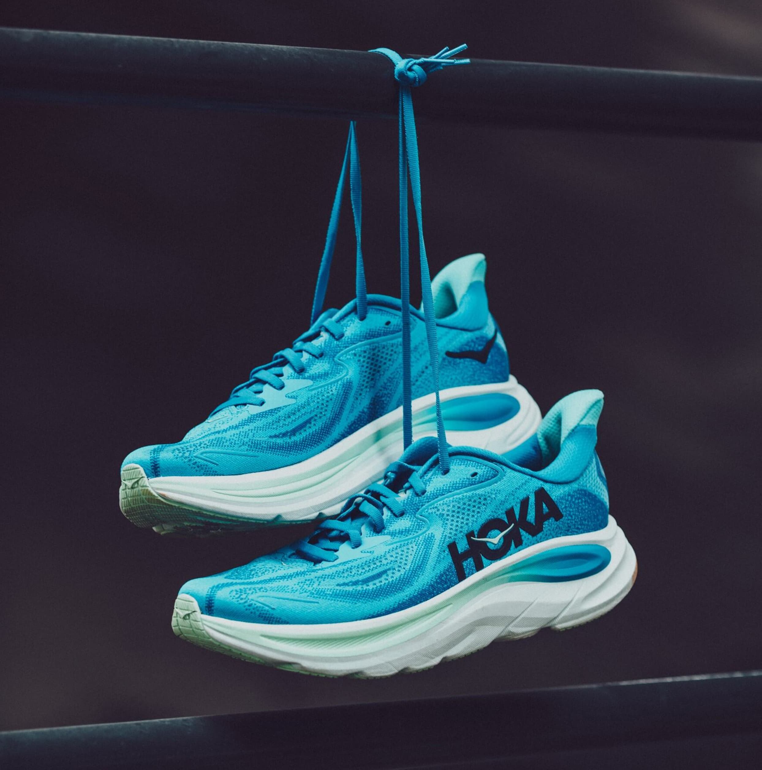 Blue running shoes with white soles hang by their laces on a black bar against a dark background.