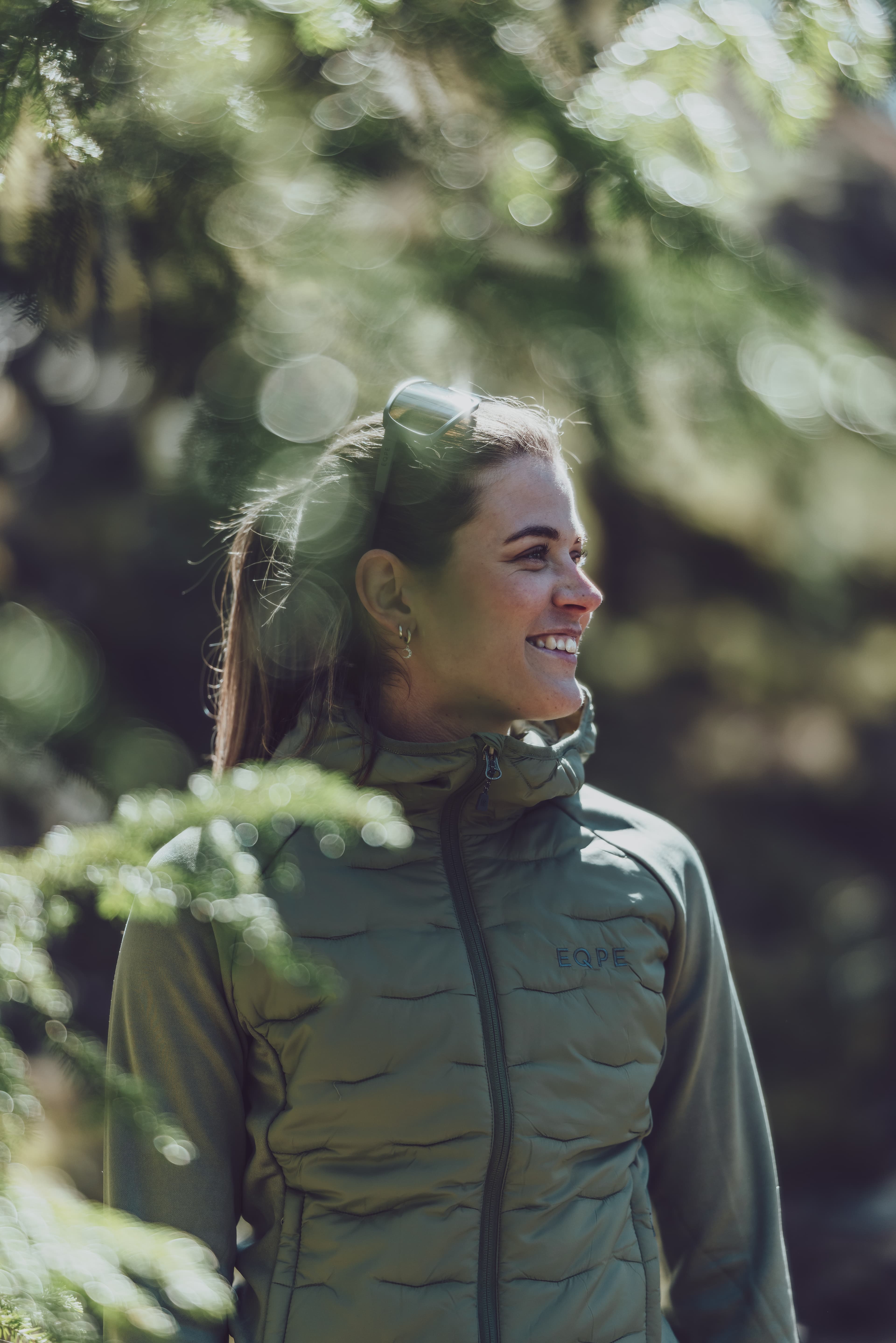 Woman in a green jacket smiling, standing outdoors surrounded by blurred greenery and sunlight filtering through trees.