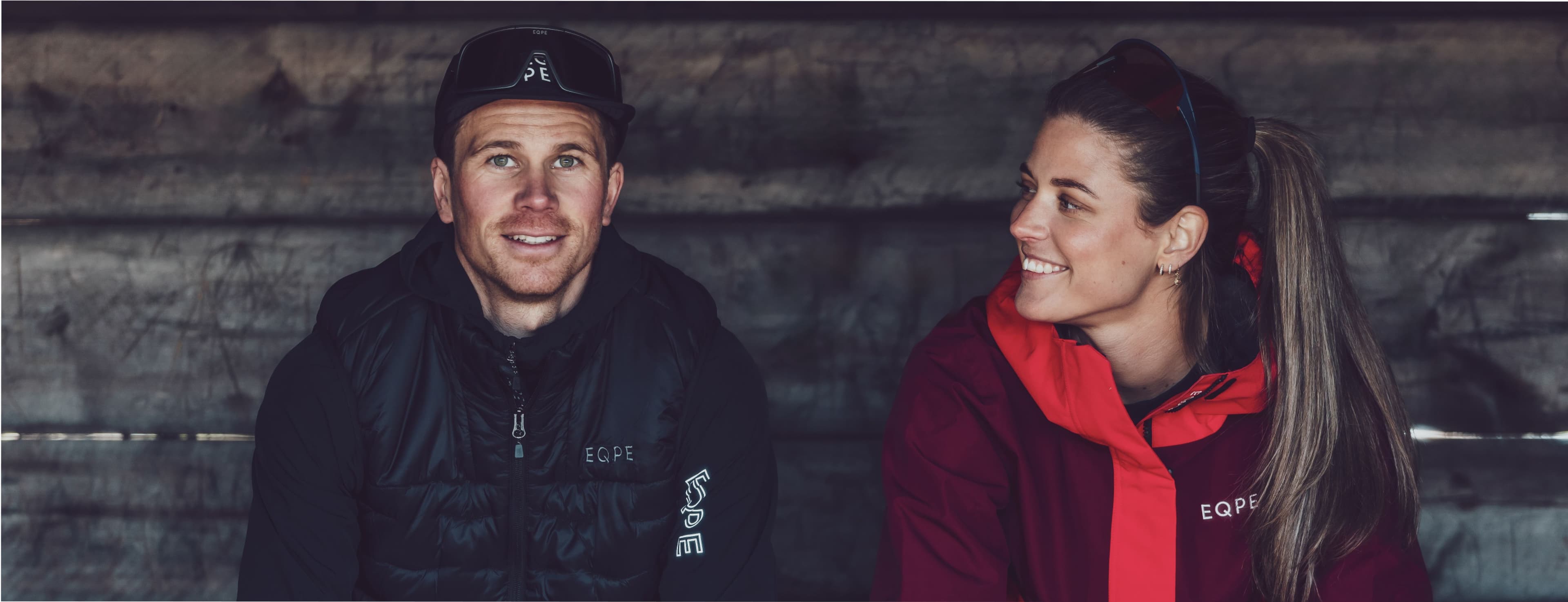 A man and woman in outdoor gear sit together, smiling, against a wooden background.
