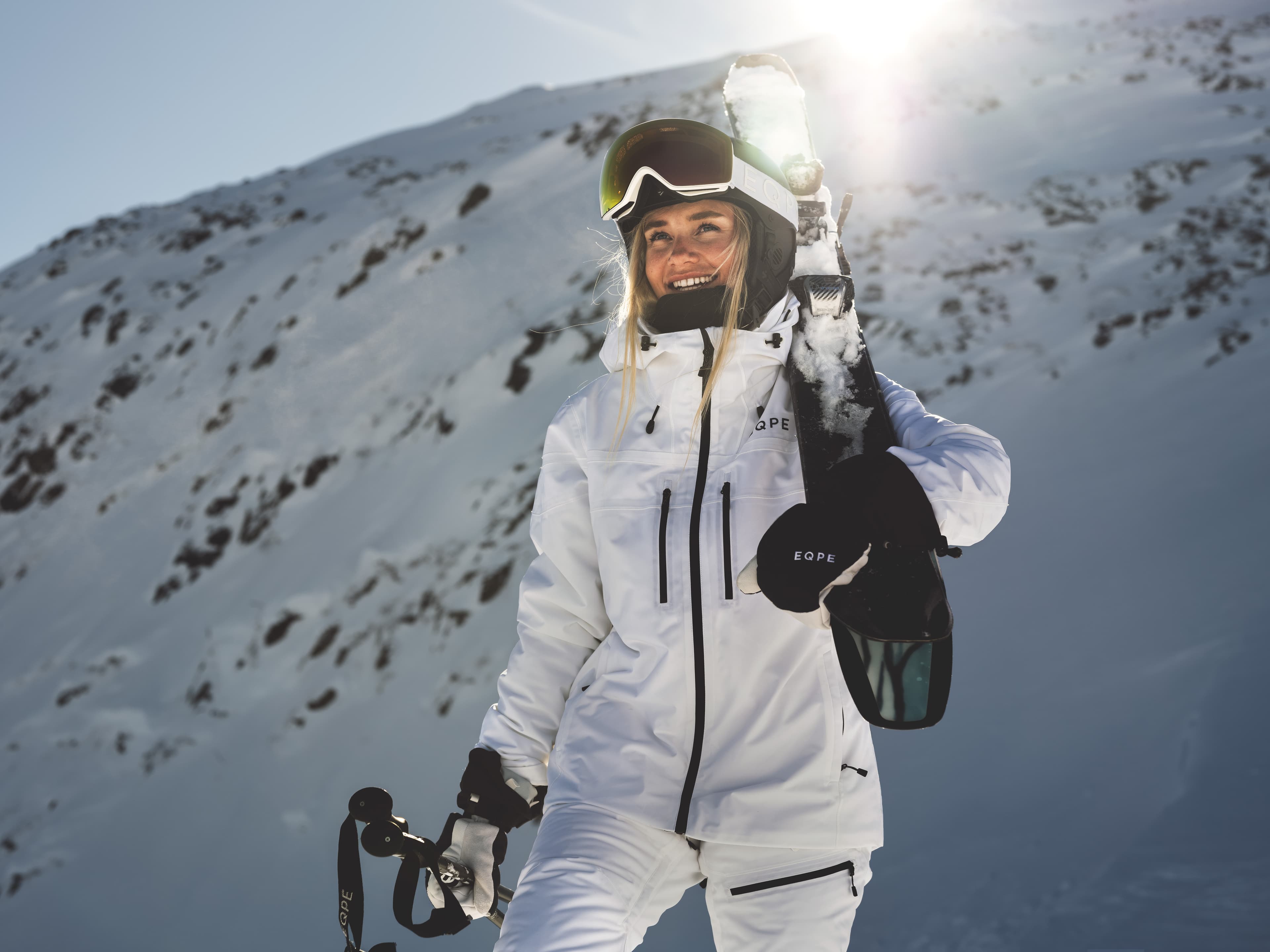 Smiling skier in white gear holds skis and poles, standing on a snowy mountain with sunlight in the background.