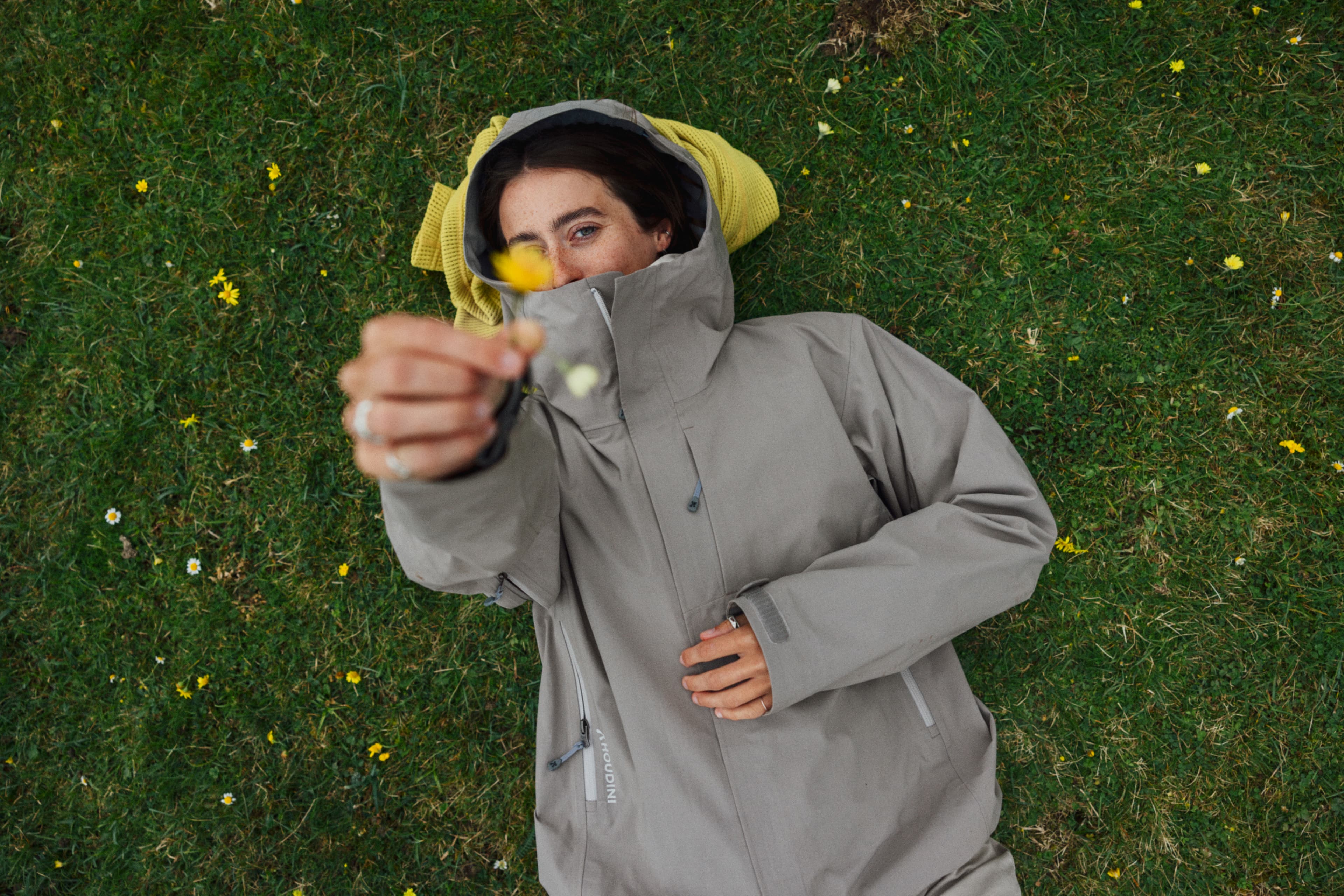 A woman lying on a field of grass wearing outdoor clothes and holding a flower